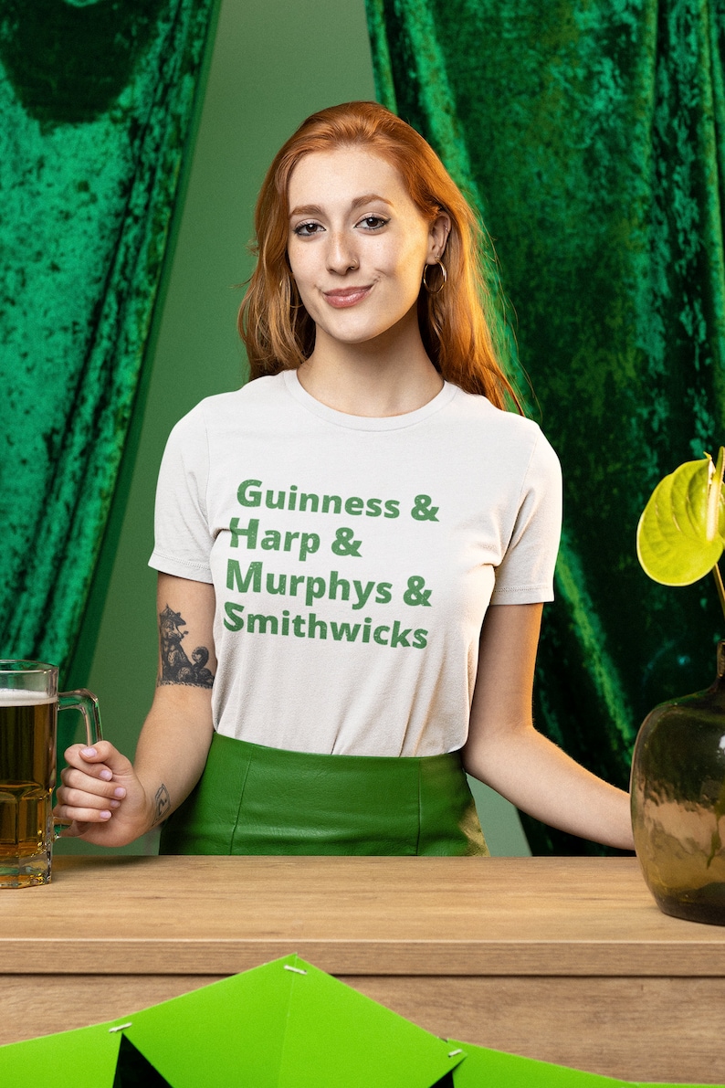 May include: A woman wearing a white t-shirt with green text that reads "Guinness & Harp & Murphys & Smithwicks" and a green skirt. She is holding a glass of beer in her right hand. The background is green.
