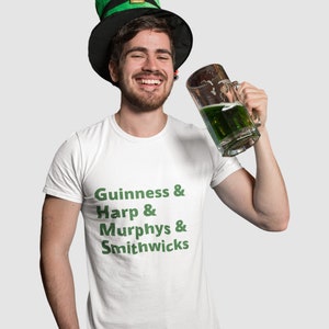 May include: A man wearing a green and black leprechaun hat is holding a glass of green beer. He is wearing a white t-shirt with the text "Guinness & Harp & Murphys & Smithwicks" printed in green.