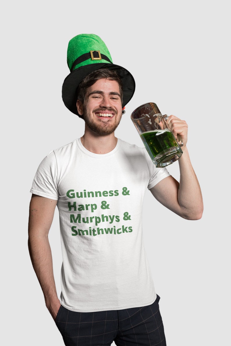 May include: A man wearing a green and black leprechaun hat is holding a glass of green beer. He is wearing a white t-shirt with the text "Guinness & Harp & Murphys & Smithwicks" printed in green.