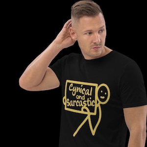 Może przedstawiać: Czarny t-shirt z napisem "Cynical and Sarcastic" w żółtym, kreskówkowym stylu. Projekt zawiera ludzika trzymającego znak. Koszulka jest noszona przez osobę.