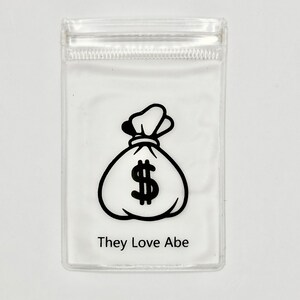 May include: A clear plastic bag with a black and white illustration of a money bag with a dollar sign. The text "They Love Abe" is printed below the illustration.