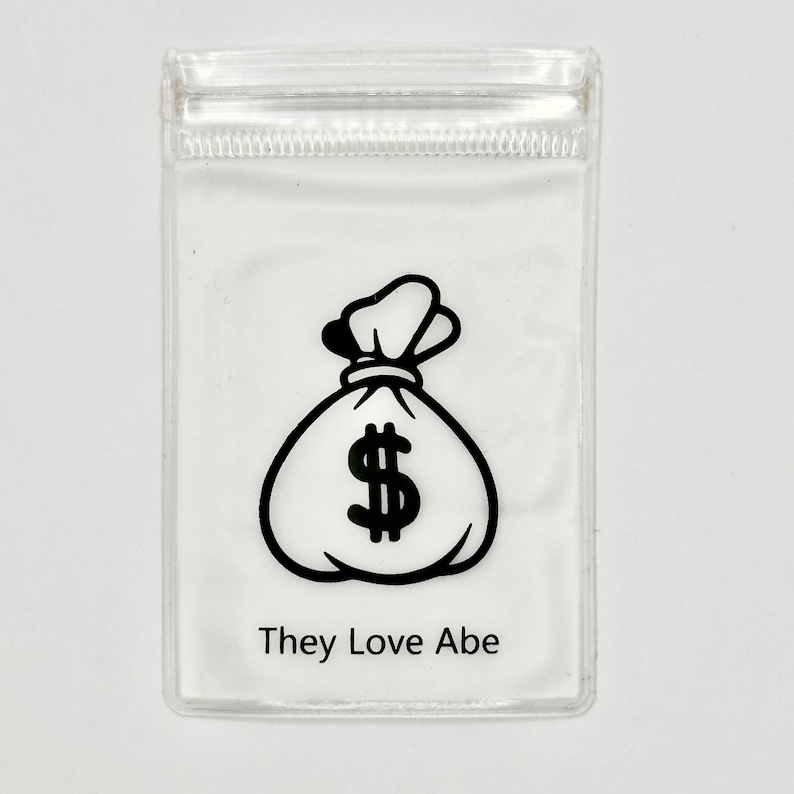 May include: A clear plastic bag with a black and white illustration of a money bag with a dollar sign. The text "They Love Abe" is printed below the illustration.