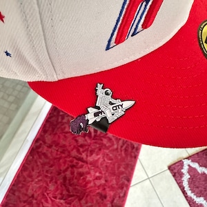 May include: A red and white baseball cap with a black and white astronaut pin. The pin features an astronaut riding a rocket with the word "CITY" on it.