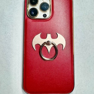 Könnte beinhalten: Rote Leder Handyhülle mit goldener Einfassung und einem goldenen Ringhalter. Die Hülle hat einen Ausschnitt in Form einer Fledermaus in der Mitte.