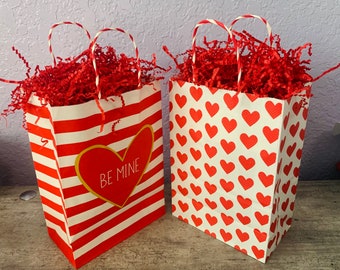 May include: Two red and white striped gift bags with red tissue paper inside. One bag has a heart with the text "Be Mine" on it. The other bag has a pattern of red hearts.