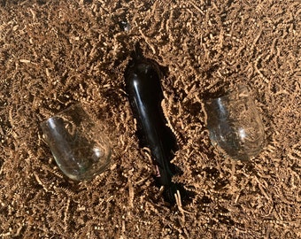 May include: A bottle of wine and two clear wine glasses are nestled in brown packing peanuts.
