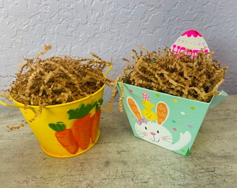 May include: Two Easter-themed containers filled with brown crinkle paper. A yellow metal bucket with carrot decorations and a green and white paper container with a bunny and chick design.