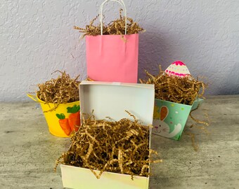 May include: Three gift boxes filled with brown shredded paper. The boxes are decorated with Easter themes. One box is pink with white handles, one is yellow with a carrot design, and one is green with a bunny design.