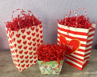 May include: Three red and white striped gift bags with red tissue paper. One bag has a heart with the text "Be Mine" on it. Another bag has a red heart pattern. The third bag is a small, floral patterned gift box.