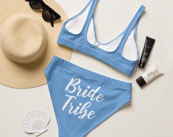 May include: A light blue bikini set with the words "Bride Tribe" printed in white on the bottoms. The set is accompanied by a straw sun hat, black sunglasses, sunscreen, and a seashell.