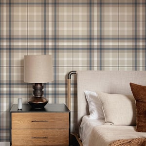 May include: A bedroom scene featuring wallpaper with a beige, white, and dark blue plaid pattern. A wooden nightstand with three drawers holds a dark brown lamp with a beige shade. A bed with a light beige headboard and textured pillows is visible.