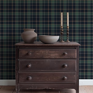 May include: A dark brown wooden dresser with three drawers, topped with a gray ceramic vase, a gray bowl, and two brass candle holders with lit candles. The background features a dark green and navy plaid wallpaper.