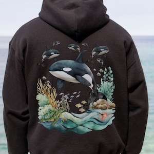 Puede incluir: Sudadera con capucha negra con un estampado gráfico de una orca nadando en un arrecife de coral. El diseño presenta una orca grande con orcas más pequeñas nadando a su alrededor. El arrecife de coral se representa con corales coloridos, peces y una tortuga marina.