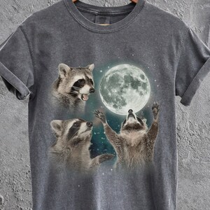 Puede incluir: Una camiseta gris oscuro con un gráfico de tres mapaches mirando hacia arriba a una luna llena. Los mapaches están en un cielo nocturno estrellado.