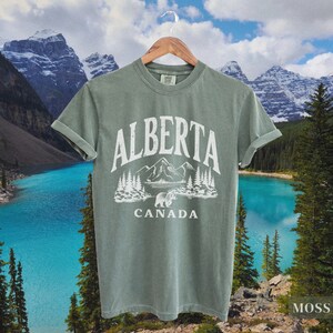 May include: A green t-shirt with a white graphic design featuring a bear, mountains, and trees. The text "ALBERTA CANADA" is printed on the shirt.