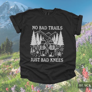 May include: Black t-shirt with white graphic of two bears hiking with backpacks, trees, and mountains. Text reads "NO BAD TRAILS JUST BAD KNEES". The word "BLACK" is in the bottom right corner.