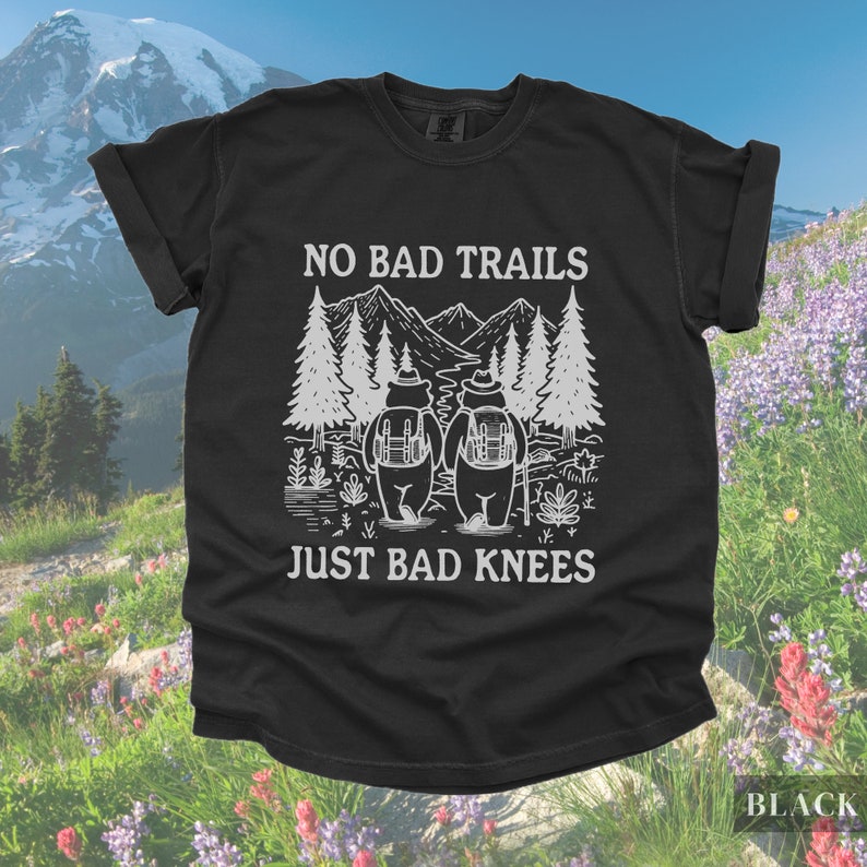 May include: Black t-shirt with white graphic of two bears hiking with backpacks, trees, and mountains. Text reads "NO BAD TRAILS JUST BAD KNEES". The word "BLACK" is in the bottom right corner.