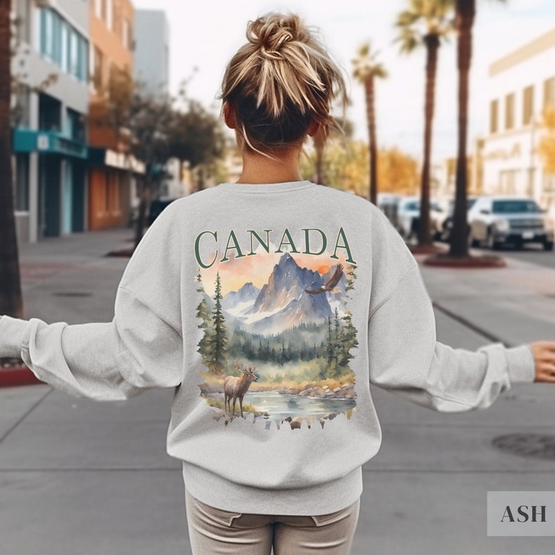 May include: A grey sweatshirt with a watercolour design on the back. The design features a mountain range, a lake, a deer, and an eagle. The word "CANADA" is printed in green above the design.