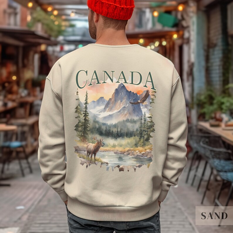 May include: A beige sweatshirt with a watercolour design of a Canadian landscape on the back. The design features mountains, a river, a deer, and an eagle. The word "CANADA" is printed in green above the design.