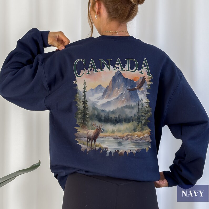 May include: Navy blue sweatshirt featuring a watercolour illustration of a Canadian landscape with mountains, a lake, a deer and a flying eagle. The word "CANADA" is printed in white above the illustration.