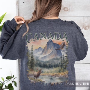 May include: Dark heather grey sweatshirt with a watercolour design of a Canadian landscape featuring mountains, a lake, a deer, and an eagle. The word "CANADA" is printed in green above the design.