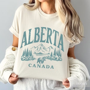 May include: A white t-shirt with a blue graphic design that says "ALBERTA CANADA" with a mountain range and a bear.