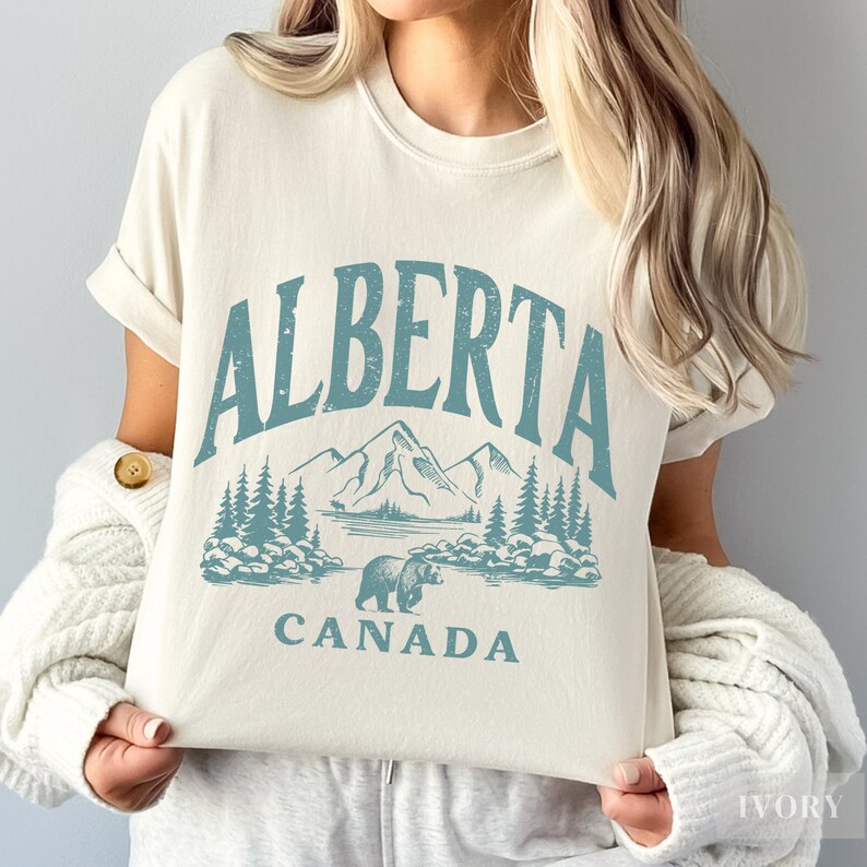May include: A white t-shirt with a blue graphic design that says "ALBERTA CANADA" with a mountain range and a bear.