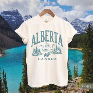 May include: A white t-shirt with a vintage-style graphic design featuring the text "ALBERTA CANADA" and a stylized image of mountains, trees, and a bear. The design is in shades of blue and green.