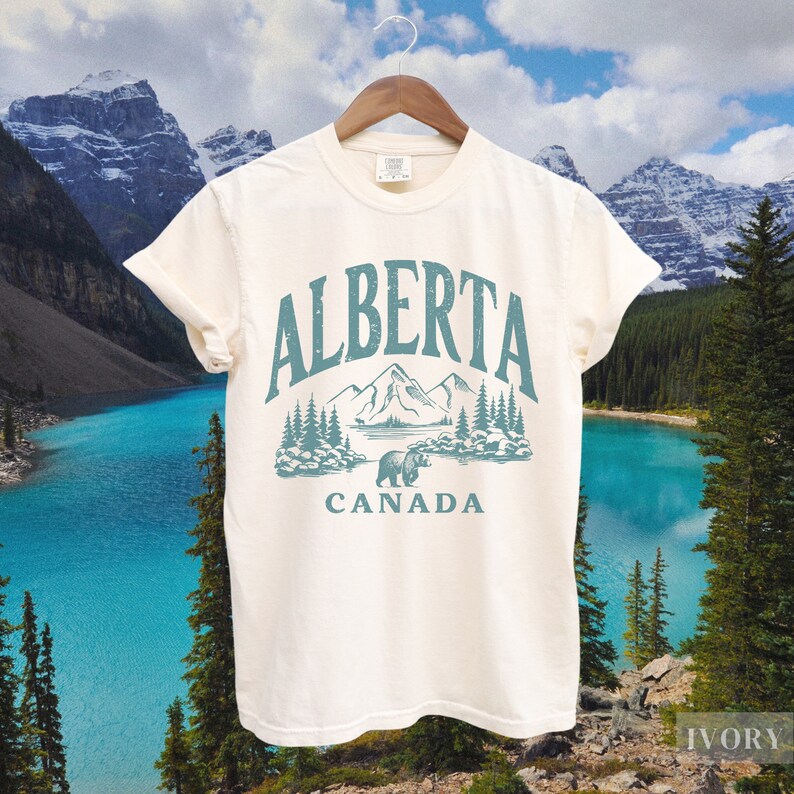 May include: A white t-shirt with a vintage-style graphic design featuring the text "ALBERTA CANADA" and a stylized image of mountains, trees, and a bear. The design is in shades of blue and green.