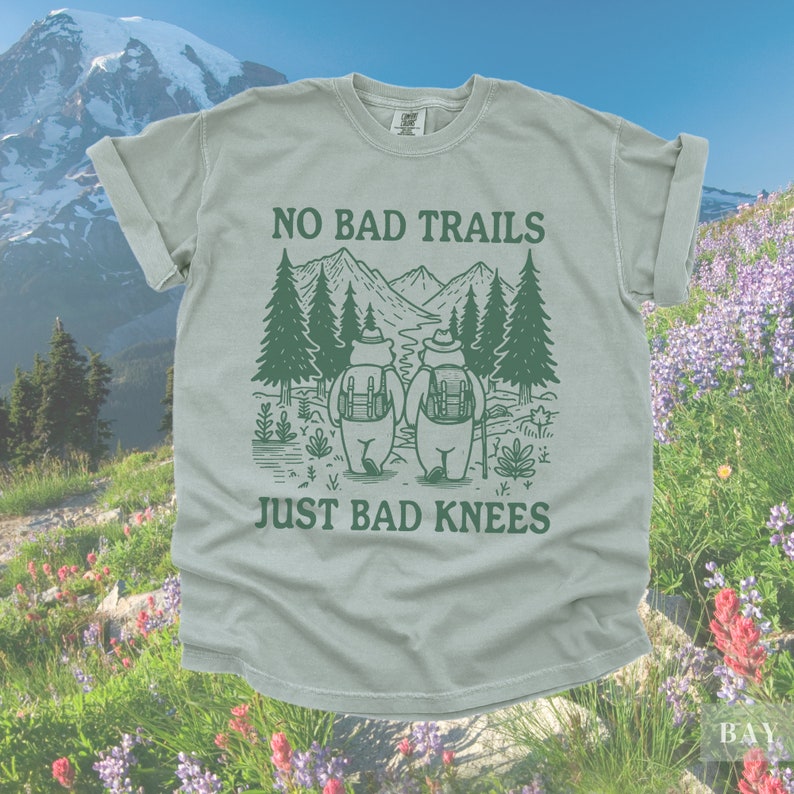 May include: A light green t-shirt with the text "NO BAD TRAILS JUST BAD KNEES" and a graphic of two bears hiking in the mountains. The bears are wearing backpacks and hats. The shirt is a comfortable, casual style.