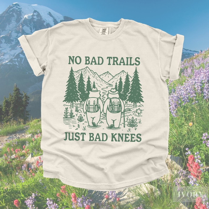 May include: Ivory-colored t-shirt with the text "NO BAD TRAILS JUST BAD KNEES" in green. The graphic features two bears with backpacks hiking in a forest with mountains in the background. The shirt has rolled-up sleeves.