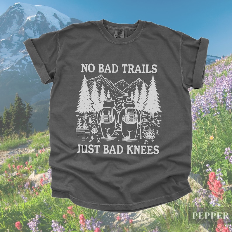 May include: Dark gray t-shirt with the text "NO BAD TRAILS JUST BAD KNEES" in white. The graphic features two bears with backpacks in front of a mountain and trees. The shirt is a Pepper color.