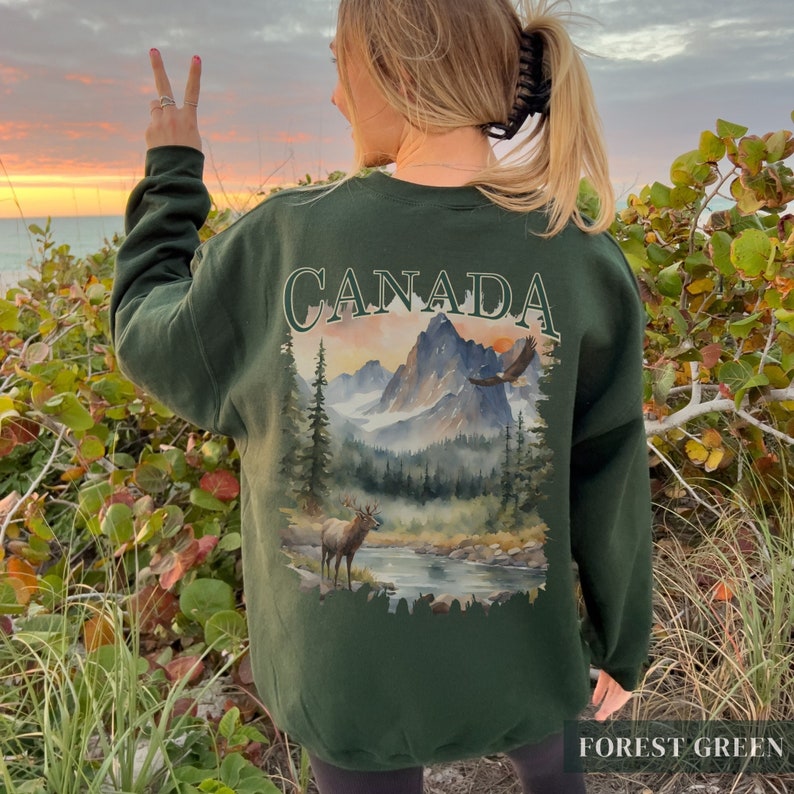 May include: A forest green sweatshirt featuring a watercolour design of a mountain range, a lake, a deer, and an eagle. The text "CANADA" is printed in white above the design. The text "FOREST GREEN" is printed in white at the bottom of the sweatshirt.