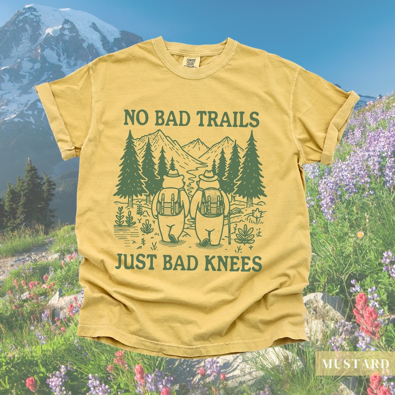 May include: Mustard-colored t-shirt with a green graphic featuring two bears with backpacks hiking in a mountain scene. The text reads "NO BAD TRAILS JUST BAD KNEES". The shirt has rolled-up sleeves and a relaxed fit.