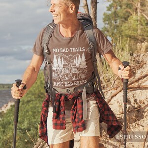 May include: Espresso-colored t-shirt with the text "NO BAD TRAILS JUST BAD KNEES" and a graphic of mountains and trees. The person is wearing a backpack, shorts, and a red and black plaid shirt tied around the waist. They are using hiking poles.