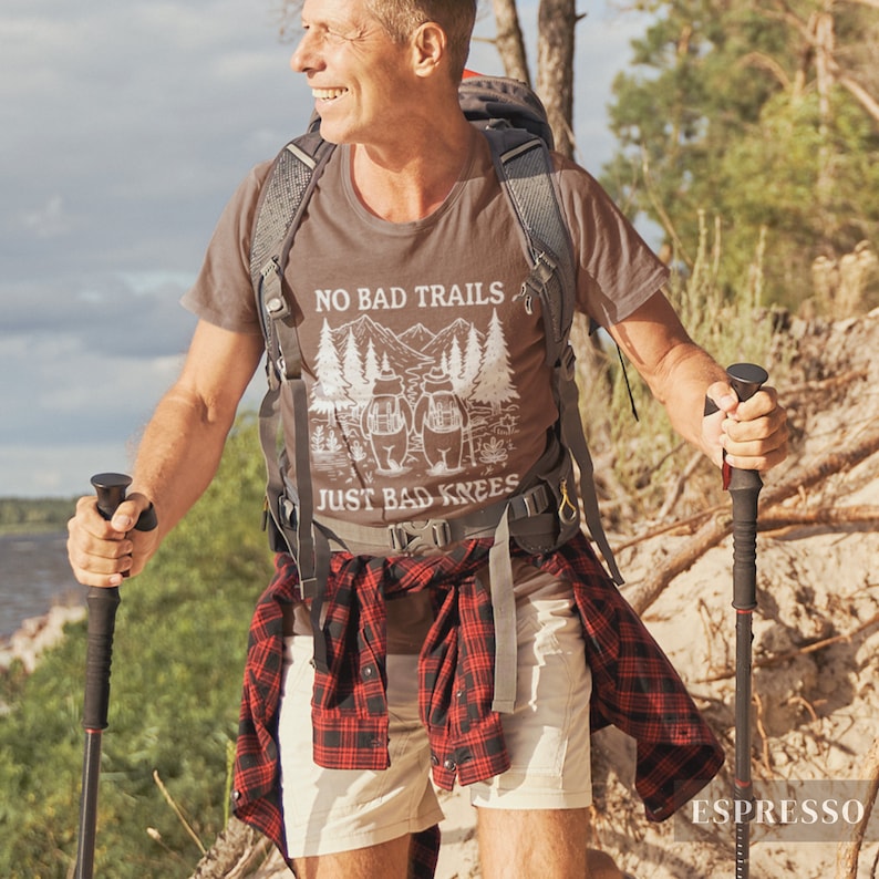May include: Espresso-colored t-shirt with the text "NO BAD TRAILS JUST BAD KNEES" and a graphic of mountains and trees. The person is wearing a backpack, shorts, and a red and black plaid shirt tied around the waist. They are using hiking poles.