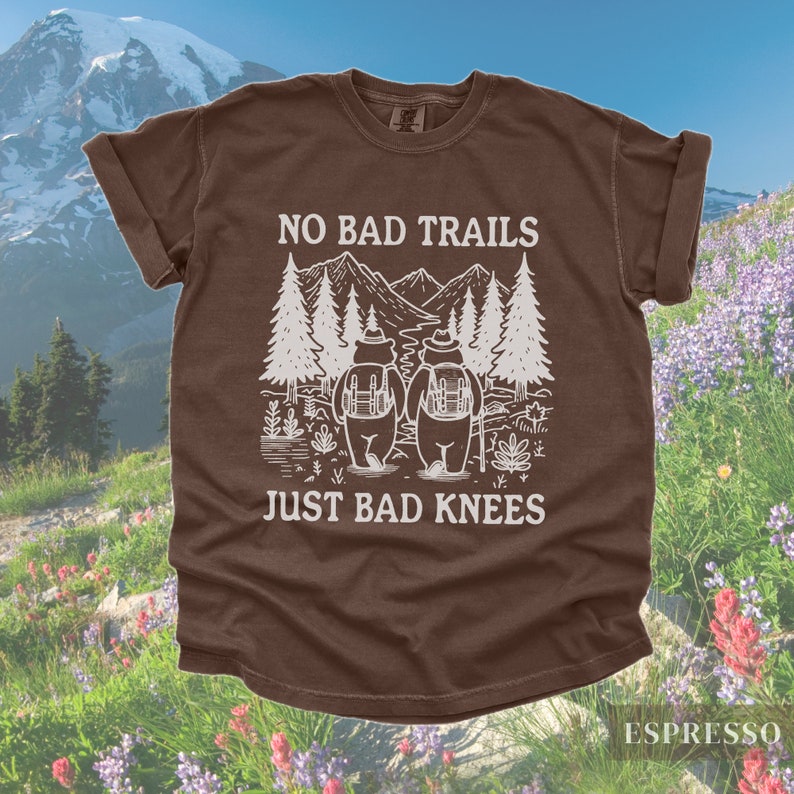 May include: Espresso-colored t-shirt with the text "NO BAD TRAILS JUST BAD KNEES" in white. The graphic features two bears with backpacks, hiking in front of mountains and trees. The shirt has rolled-up sleeves.