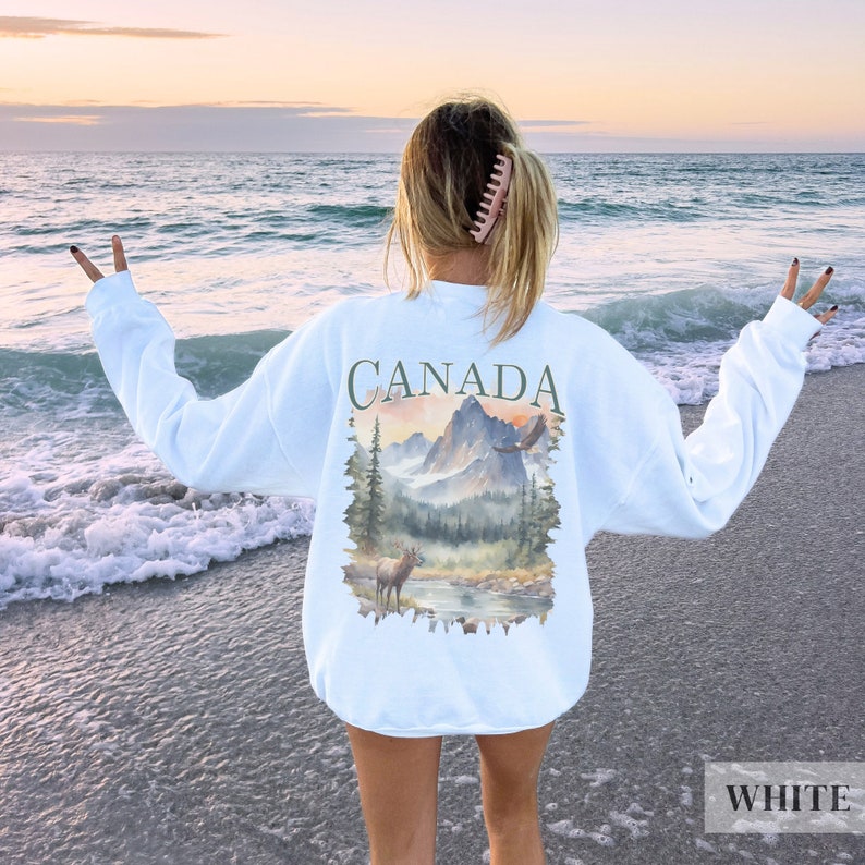 May include: White sweatshirt with a graphic print of a mountain scene with a deer and an eagle. The text "CANADA" is printed above the graphic.