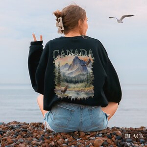 May include: Black sweatshirt featuring a graphic print of a mountain landscape with a deer and an eagle. The text "CANADA" is printed in green on the sweatshirt.