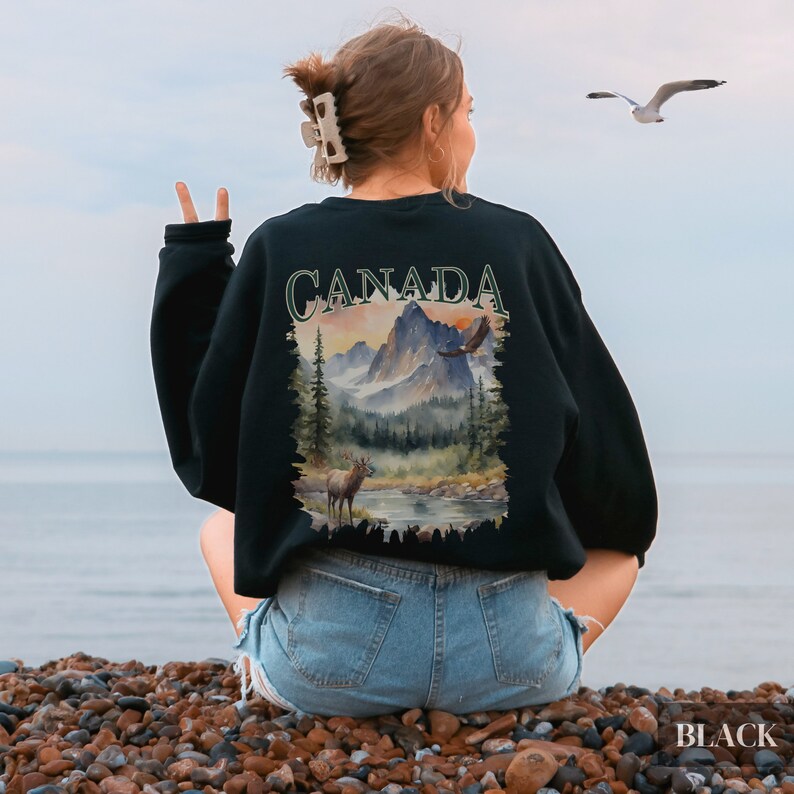 May include: Black sweatshirt featuring a graphic print of a mountain landscape with a deer and an eagle. The text "CANADA" is printed in green on the sweatshirt.