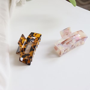Könnte beinhalten: Zwei rechteckige Haarspangen. Eine Spange hat ein dunkelbraunes und schwarzes Schildpattmuster mit glänzender Oberfläche. Die zweite Spange besteht aus einem durchscheinenden, perlmuttweißen Material mit dezenten rosa und irisierenden Flecken.