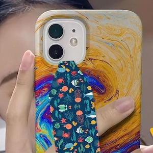 May include: A phone case with a colorful abstract design and a blue phone grip with a sea life pattern. The grip has a variety of sea creatures including fish, crabs, starfish, and seahorses.