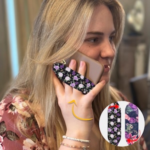 May include: A purple and black floral phone grip with a black background. The grip is designed to be placed on the back of a phone and used to hold the phone more securely. The grip is shown being used on a phone, with the user's hand holding the phone to their ear.
