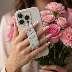 May include: A light beige phone case with a patterned strap is held in front of a bouquet of pink roses and carnations. The phone case has a camera bump with three lenses. The strap has a white background with green and yellow floral details.