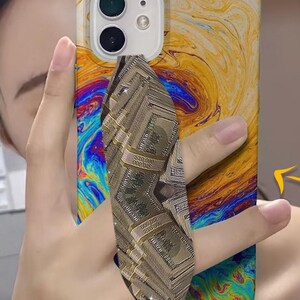 May include: A phone case with a colorful abstract design featuring swirling colors of blue, green, yellow, and orange. A phone grip with a stack of money design is attached to the back of the case.