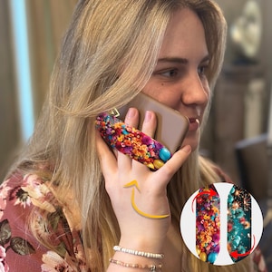 May include: A woman is holding a phone with a colorful floral phone grip attached to the back. The grip is designed to help people hold their phones more securely. The grip is also a stylish accessory that can add a touch of personality to any phone.