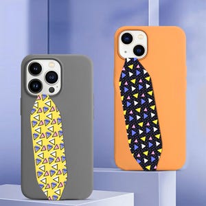 May include: Two smartphone cases, one matte gray and one matte orange, each featuring a vertical strap with a geometric pattern. The gray case has a yellow strap with blue, pink, and white triangles, while the orange case has a black strap with similar colorful triangles.