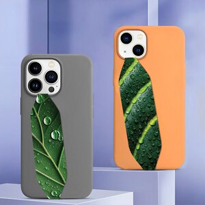 May include: Two phone cases, one grey and one orange, each feature a vibrant green leaf design with water droplets. The leaf design is a vertical accent on the back of the phone cases. The cases are made of a smooth material.