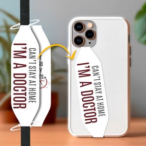 May include: White phone case with a white strap attached. The strap has the text "Can't stay at home I'm a doctor" printed on it in maroon.