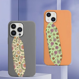 May include: Two smartphone cases, one matte gray and one matte orange, each featuring a vertical, leaf-shaped adhesive strap. The gray case has a cream-colored strap with a pink and orange floral pattern, while the orange case has a light green strap with a pink and orange floral pattern.